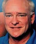 A man with white hair and blue shirt smiling.